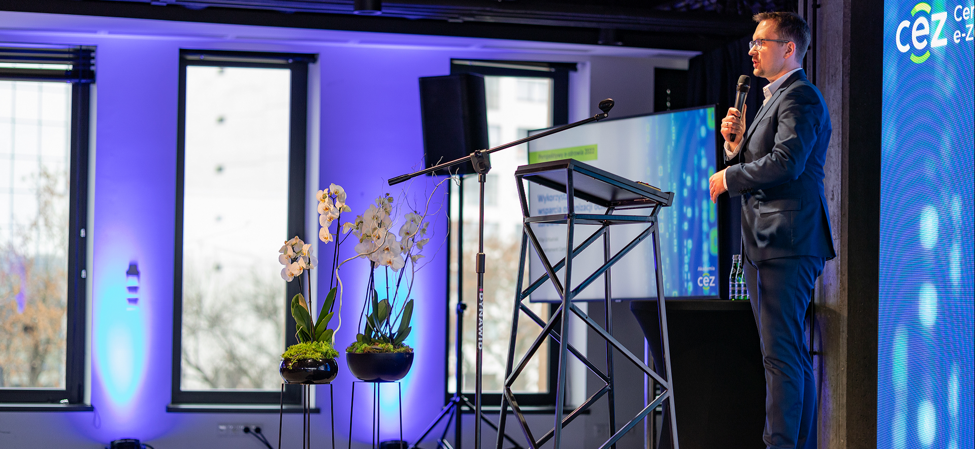 zdjęcie przedstawia stojącego przy pulpicie mężczyznę, który w ręku trzyma mikrofon. Za jego plecami jest wyświetlana prezentacja