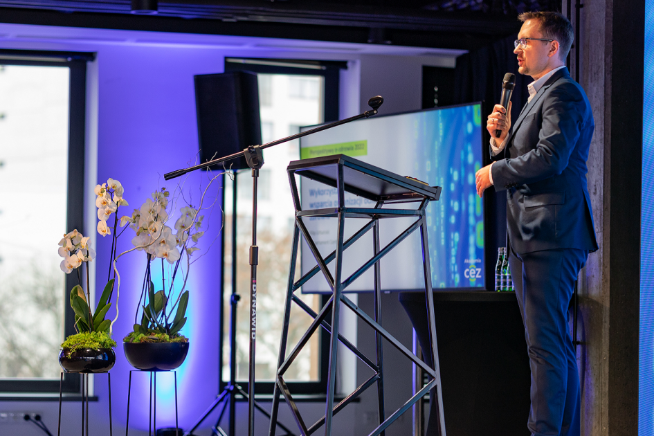 zdjęcie przedstawia stojącego przy pulpicie mężczyznę, który w ręku trzyma mikrofon. Za jego plecami jest wyświetlana prezentacja